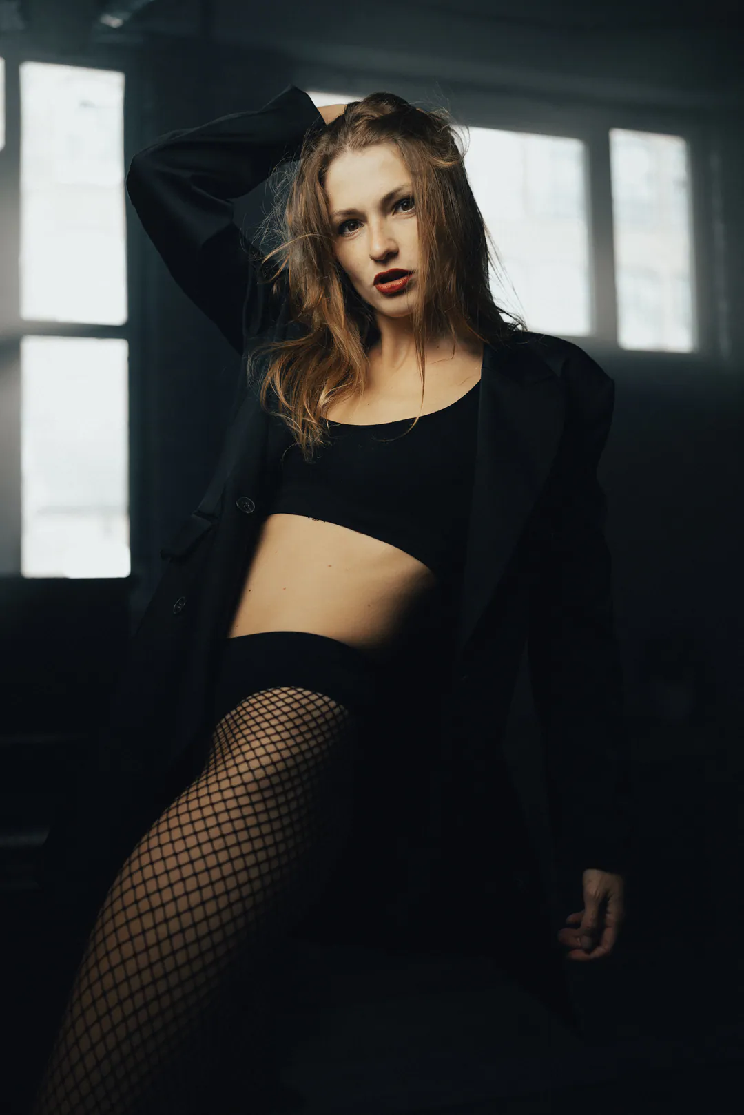Woman in a black crop top, long jacket and fishnet tights, hand in hair, in a dark room