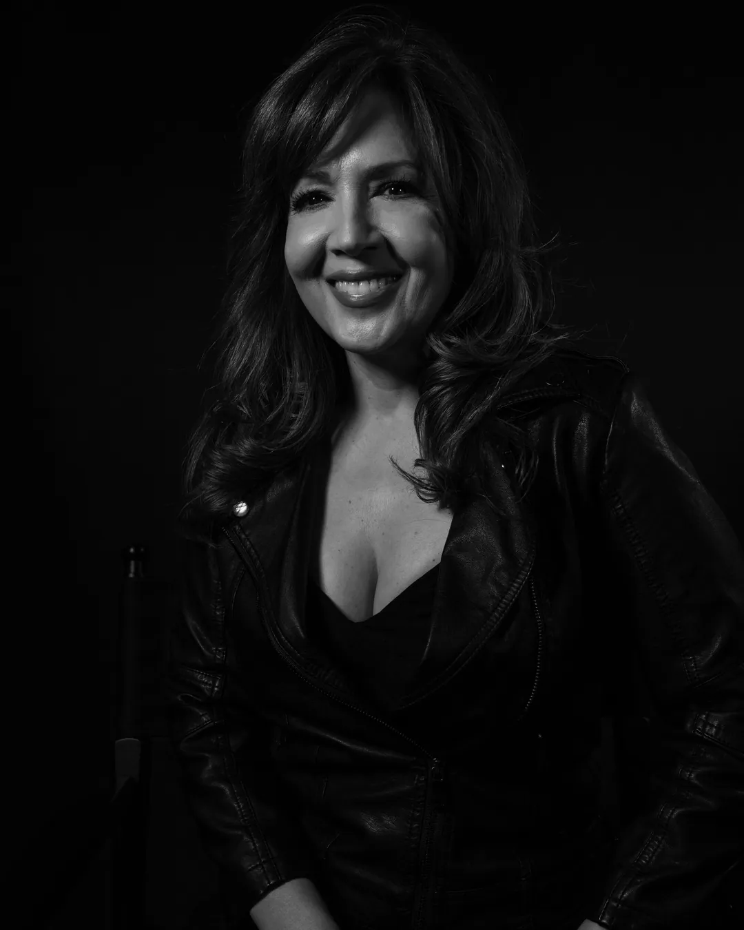 Woman with long wavy hair in a leather jacket smiling softly against a dark backdrop, black and white