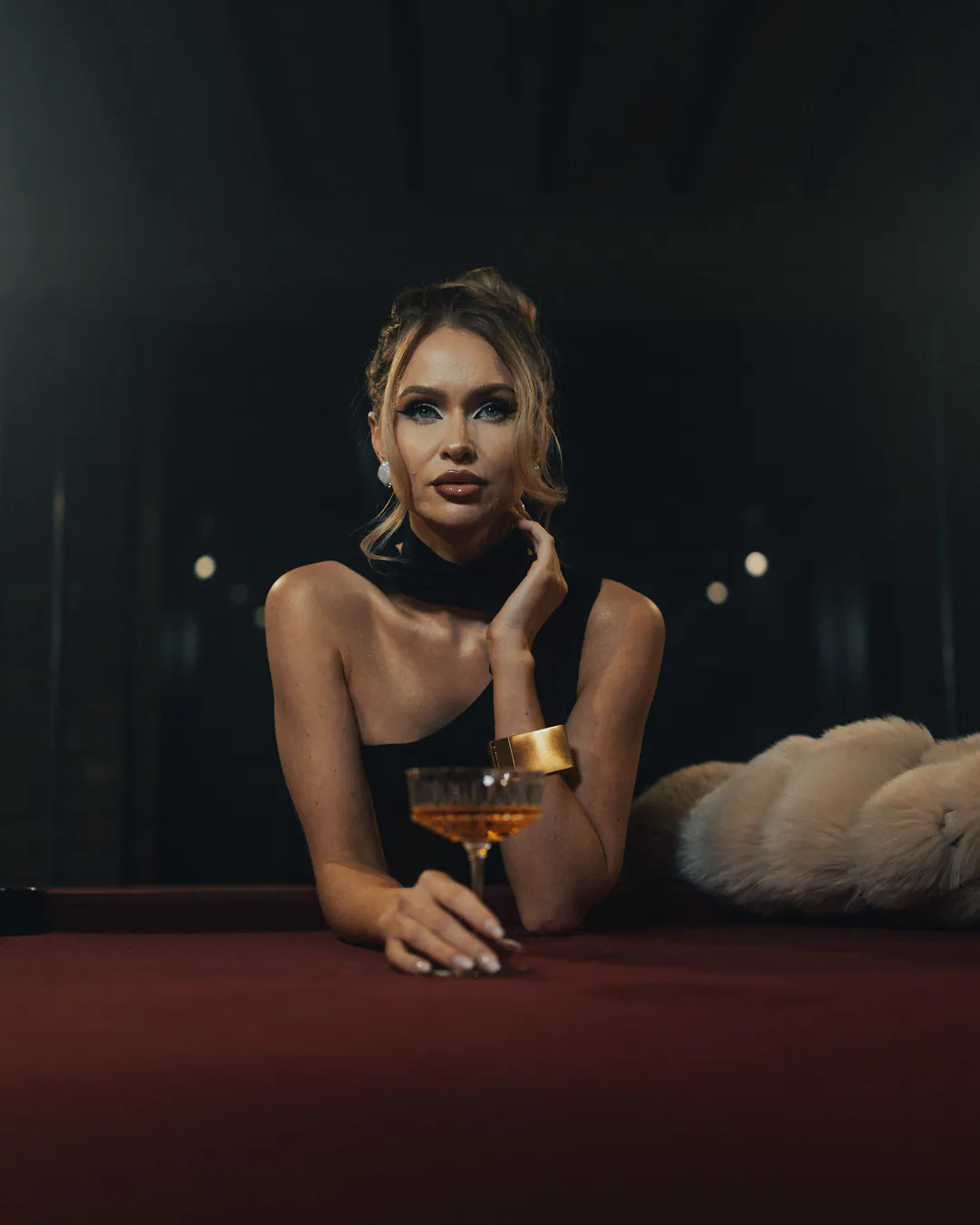 Woman in a black one-shoulder top leaning on a red baize table, holding a coupe glass