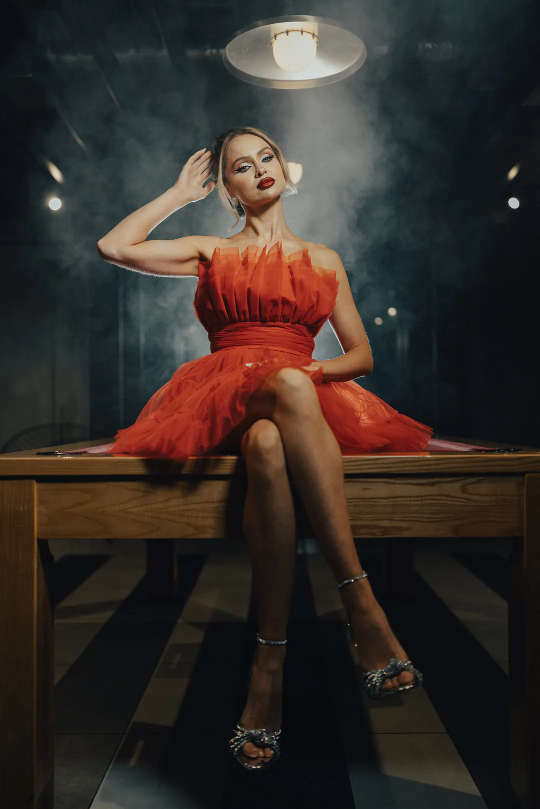 Woman in a red ruffled tulle dress sitting cross-legged on a wooden table in smoky darkness