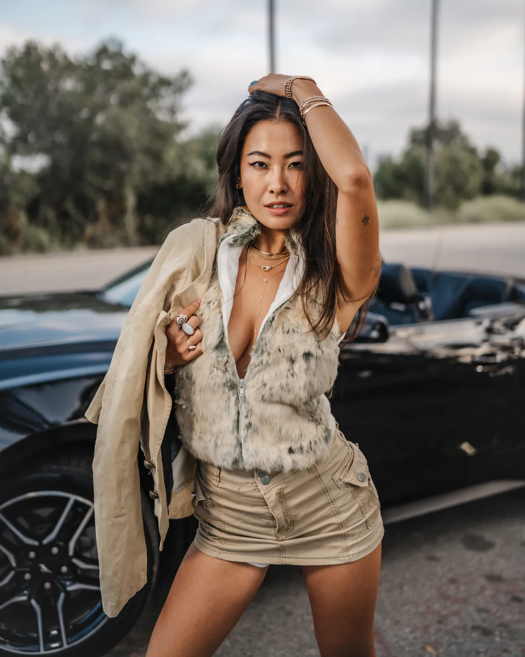 Woman in a faux fur waistcoat and khaki mini skirt, jacket over her shoulder, beside a black convertible