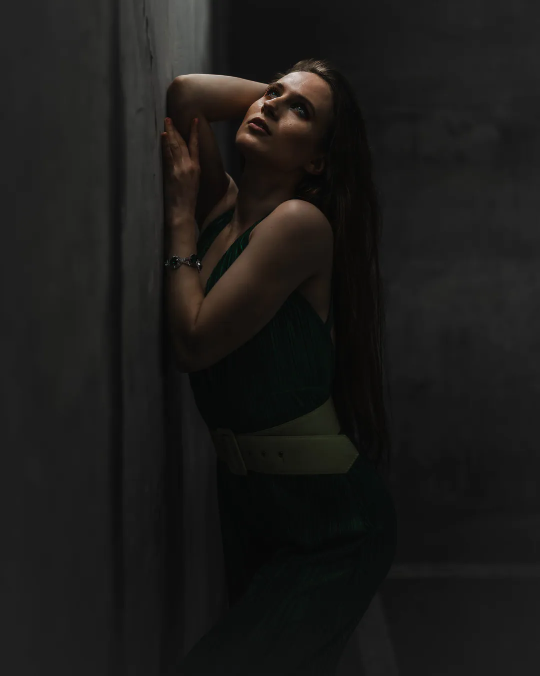 Woman in a green top and pale belt leaning against a concrete wall, looking upwards in low light