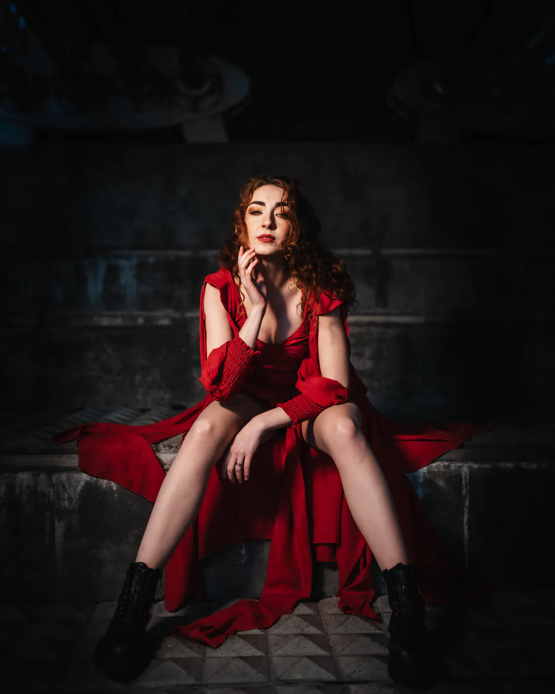 Woman in a flowing red dress and black boots seated on a stone ledge against dark brickwork