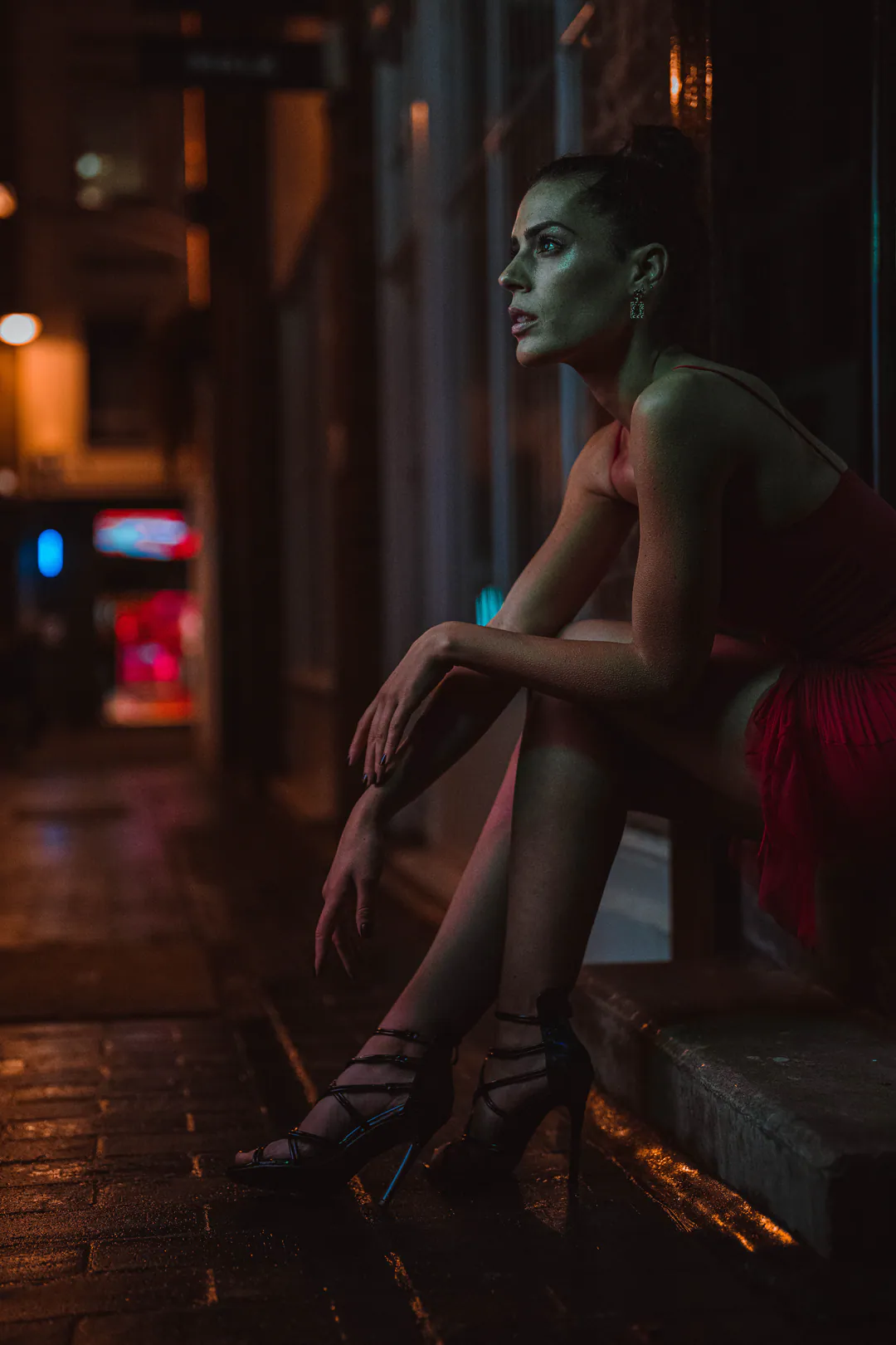 Woman in a red dress sitting on a doorstep at night, gazing down a glowing city street