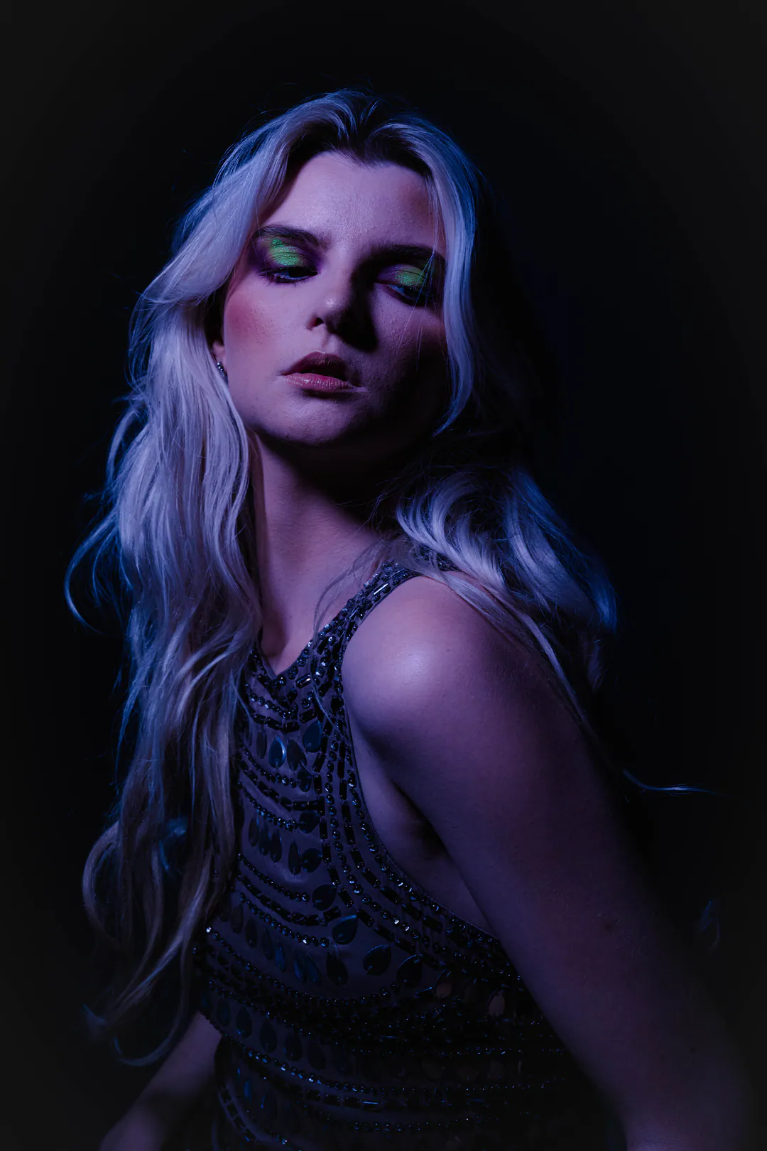 Woman with long wavy hair in an embellished top, eyes lowered, lit in violet and blue tones