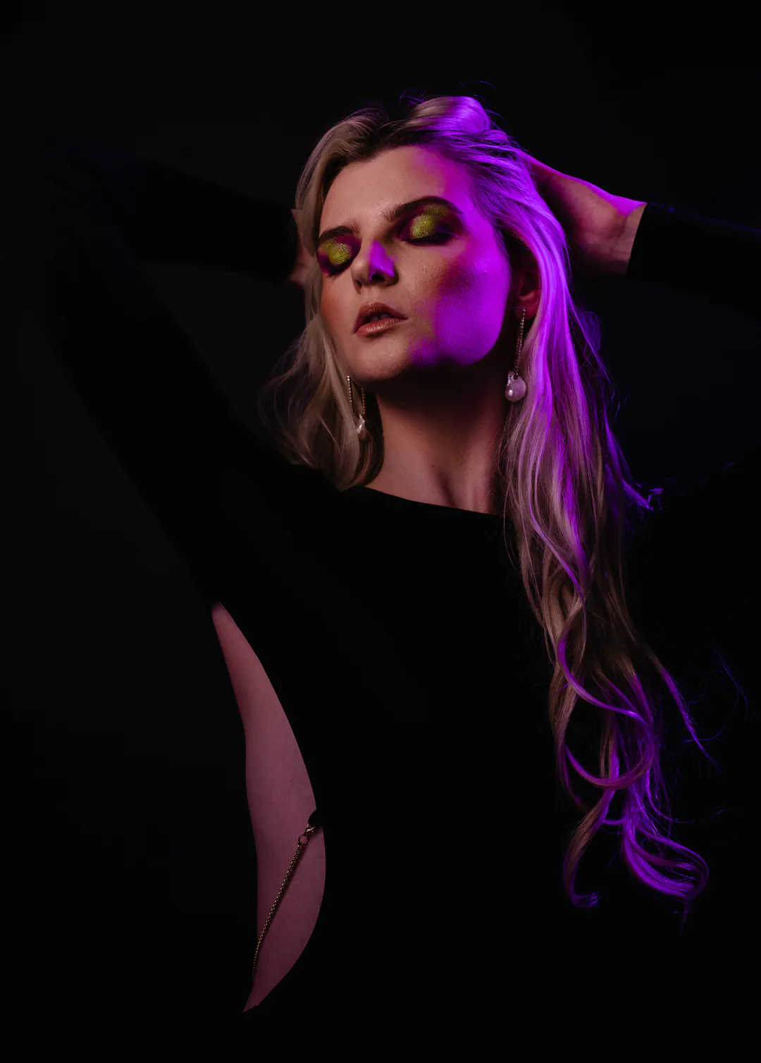 Woman with long hair and a hand behind her head, eyes closed, lit by magenta light on black