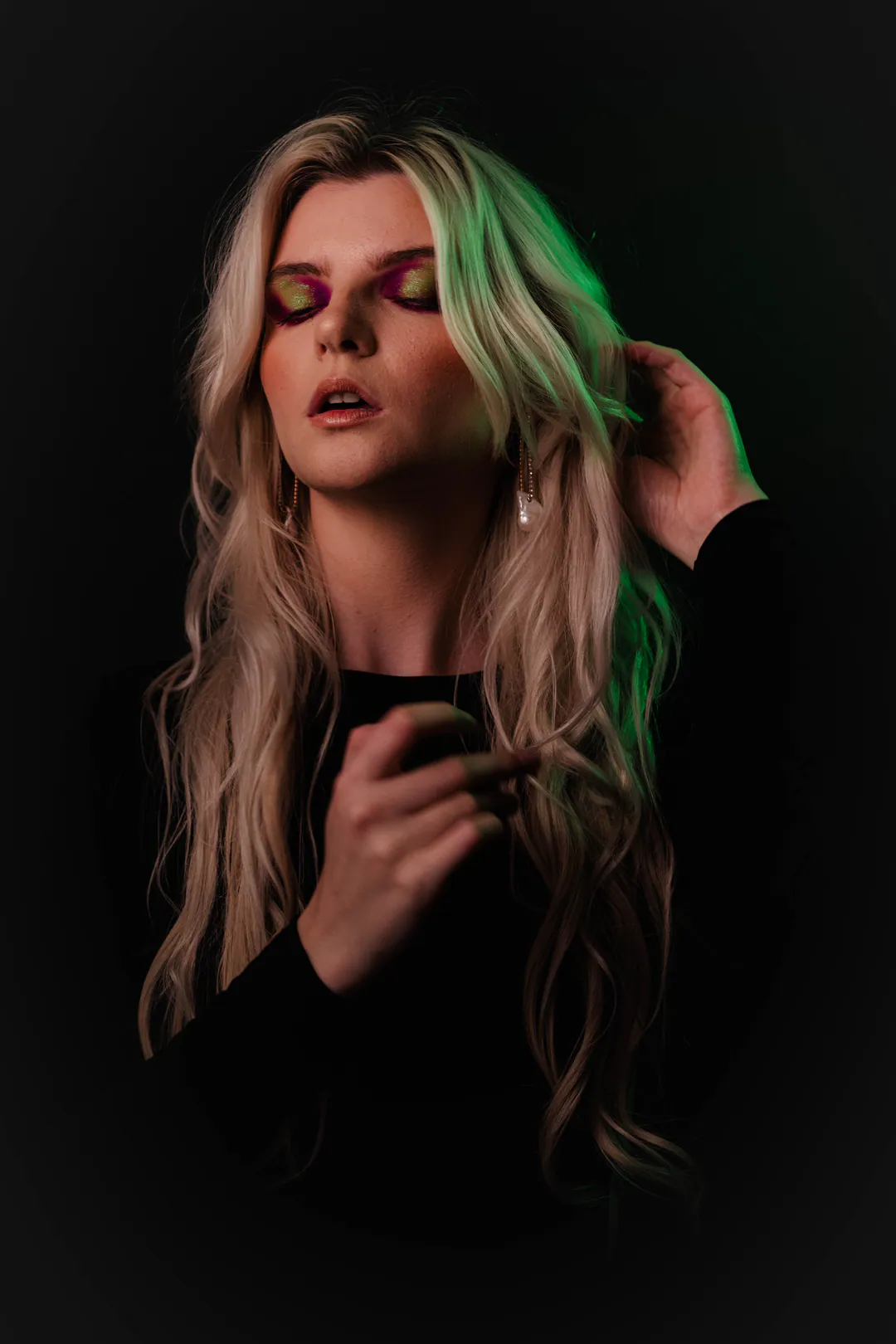 Woman with long blonde waves touching her hair, eyes closed, under green and pink coloured light