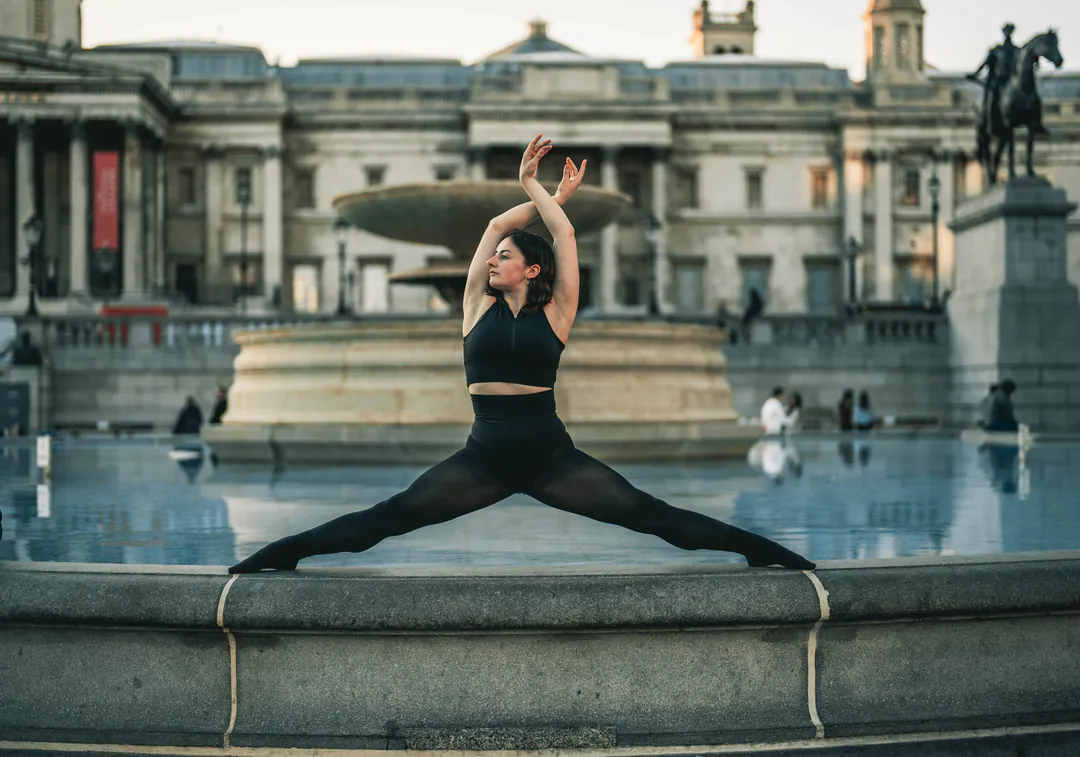 Dancer in a black crop top and leggings doing the splits on a fountain edge in Trafalgar Square