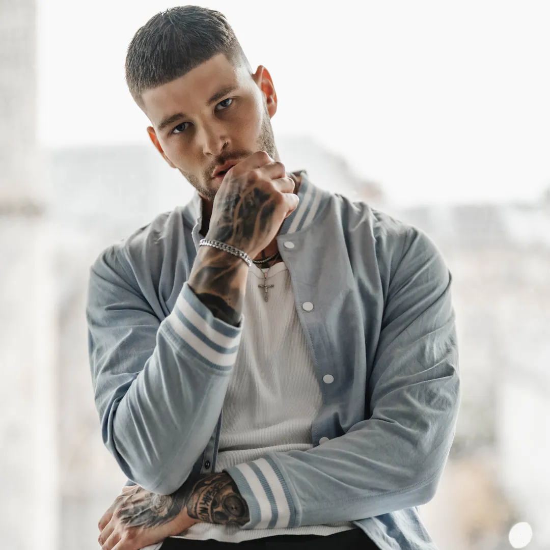 Tattooed man in a pale blue varsity jacket resting his chin on his hand beside a bright window