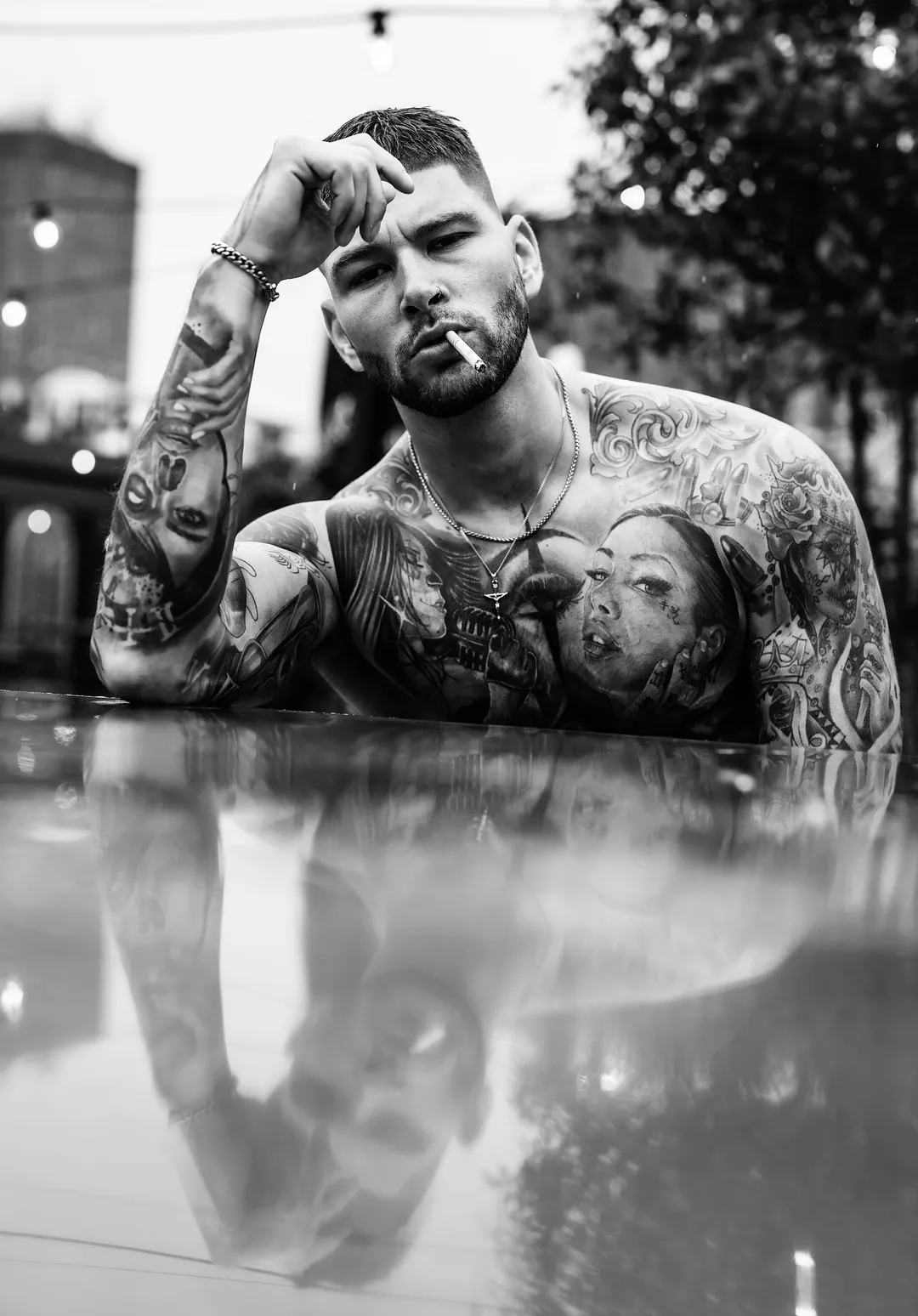 Shirtless tattooed man with a cigarette leaning on a glossy reflective surface outdoors, black and white