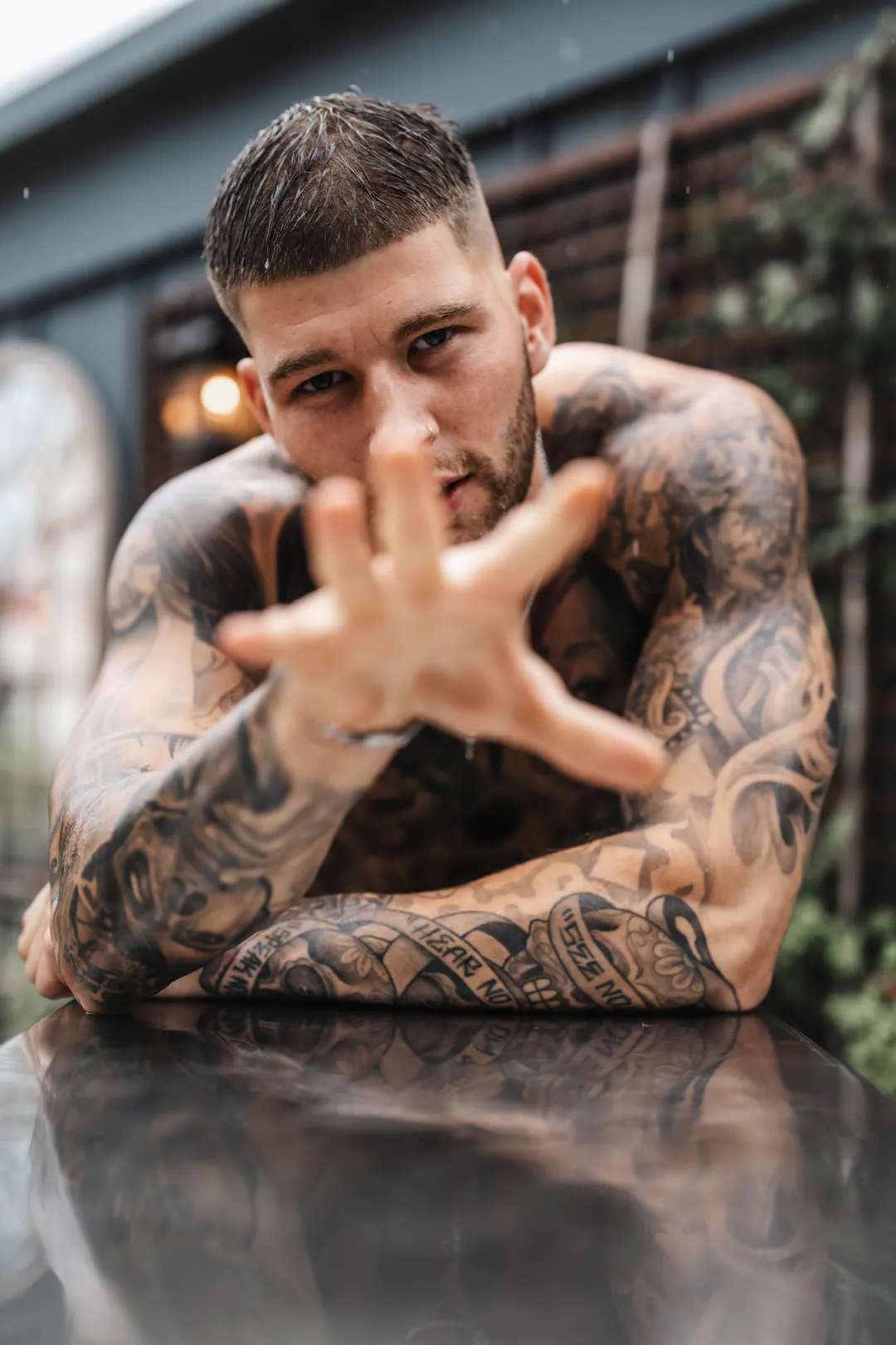 Shirtless tattooed man reaching an open hand towards the camera while leaning on a reflective surface